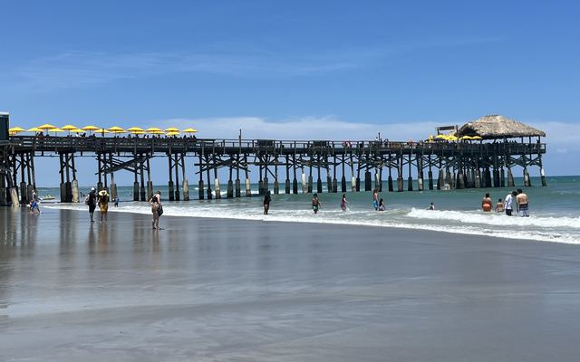 The Cocoa Beach Pier, a long wooden pier with yellow umbrellas and a thatched roof at its end, seen from the wet sand as people wade in the surf