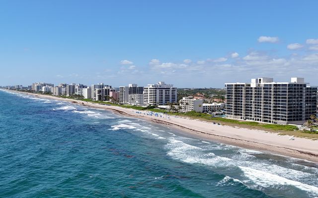 Aerial view of Highland Beach, Florida: mostly white high-rise towers set back behind a wide sandy beach, with blue-green Atlantic surf along the shore and more towers stretching into the distance