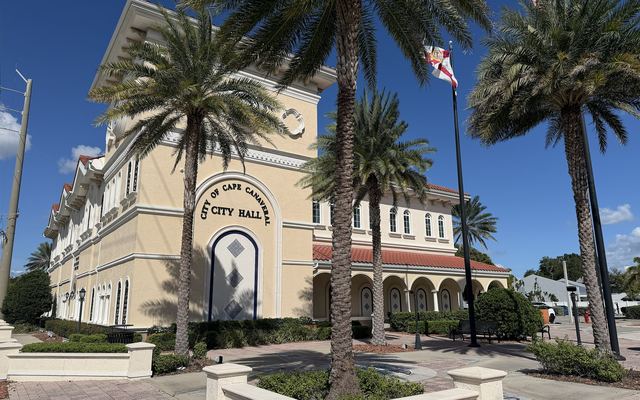 Cape Canaveral City Hall, a cream-colored building with arched windows, a red tile roof and the words City of Cape Canaveral City Hall above a tall arch, framed by tall palms and a Florida flag under a blue sky