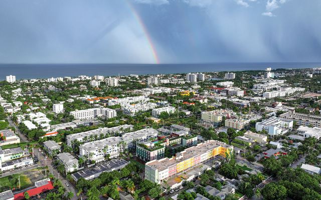Aerial view over the low-rise rooftops and palm trees of Delray Beach toward the Atlantic Ocean, with a rainbow over the water