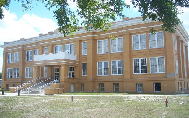 The historic Fellsmere School, a tan brick building with two floors of tall white-framed windows over a raised base and a railed front staircase, seen across a lawn under overhanging tree branches