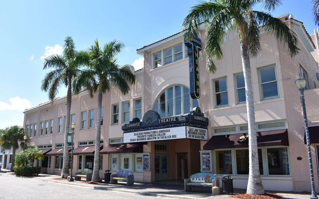 The pink stucco front of the historic Sunrise Theatre on a brick-paved street in downtown Fort Pierce, with a tall vertical Sunrise sign, a marquee over the entrance and palm trees along the sidewalk