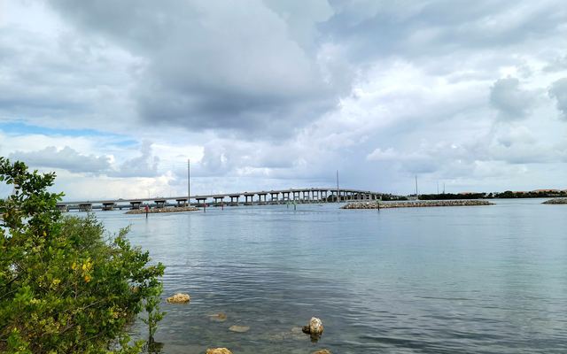 The A1A bridge to South Hutchinson Island stretching across the calm Indian River Lagoon at Fort Pierce, with rock breakwaters in the water, green shrubs and shoreline rocks in the foreground and gray clouds overhead