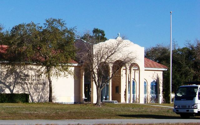 The Martin County Public Library Indiantown branch: a cream stucco building with a red tile roof, an arched entry colonnade and the branch name lettered on its front wall, behind a grass lawn under a clear blue sky