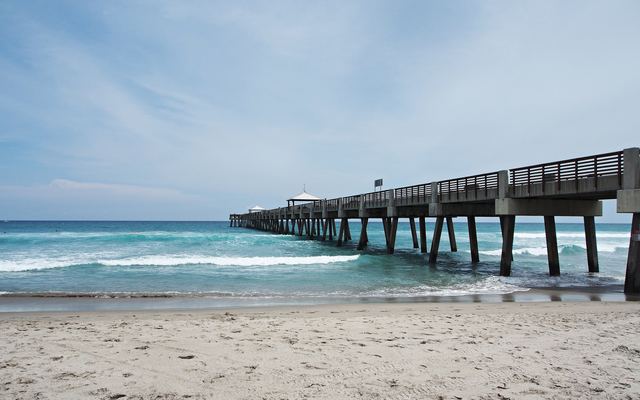 The long concrete Juno Beach Pier running from the sand out over turquoise surf, with small roofed shelters along it, under a pale cloudy sky