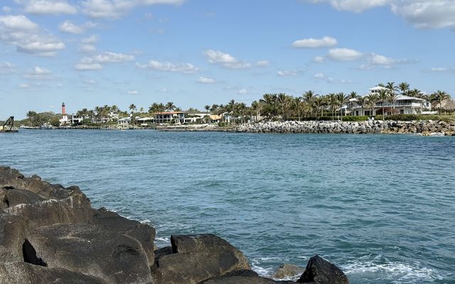 The Jupiter Inlet seen from the rocks at DuBois Park, with turquoise water, a stone jetty and waterfront homes under palm trees on the far shore, and the red Jupiter Inlet Lighthouse in the distance on the left