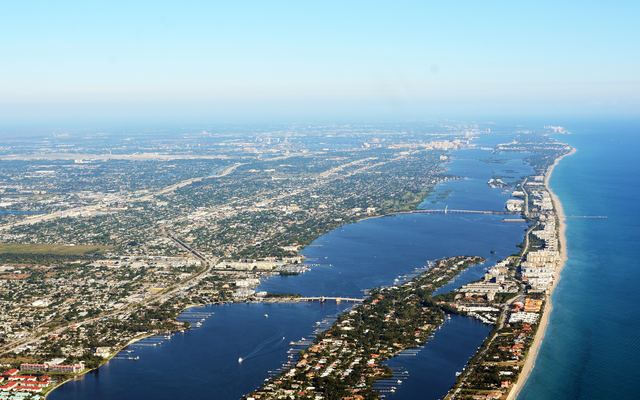 Aerial view looking north over Lantana and South Palm Beach: the Lake Worth Lagoon with two bridges to the barrier island, waterfront homes with boat docks, beachfront towers and the Atlantic shore