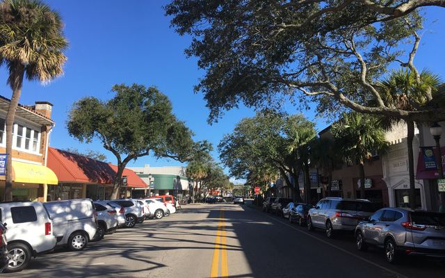 East New Haven Avenue in downtown Melbourne on a clear day, with palms and spreading live oaks shading parked cars and low storefronts on both sides of the street