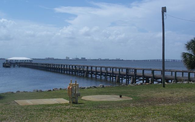 The long wooden Melbourne Beach Pier running out over the calm Indian River Lagoon to a white-roofed covered shelter at its end, seen from a grassy shore with a palm tree at the right and the low mainland skyline along the horizon