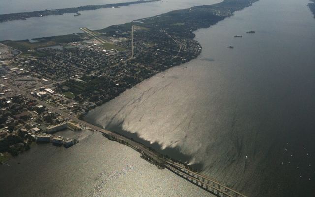 Aerial view of Merritt Island from about 4,000 feet, showing neighborhoods and the Merritt Island Airport runway, wide lagoon water on both sides of the island and a long bridge crossing the water in the foreground