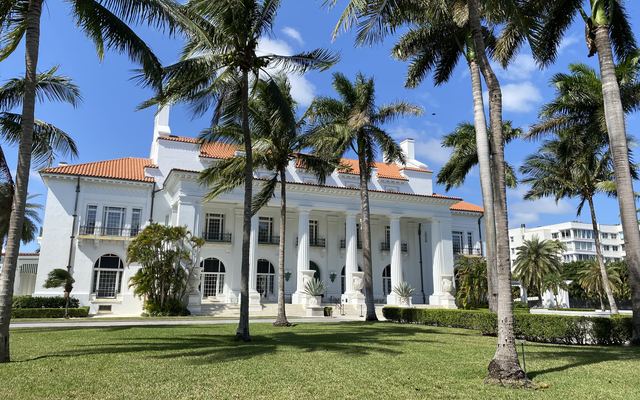 Whitehall, the white-columned Flagler Museum mansion with a red tile roof, seen across a sunny lawn dotted with tall palm trees in Palm Beach