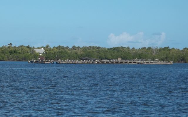 A long wooden dock along the tree-lined shore of St. Lucie Inlet Preserve State Park, seen across open blue water under a blue sky with a few clouds