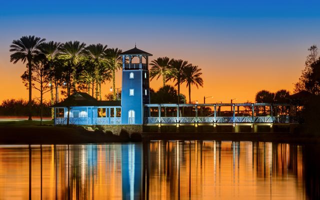 A blue lookout tower and covered pavilion with a long railed walkway on the shore of Lake Tradition in Port St. Lucie at twilight, palm trees silhouetted against an orange sky and their lights reflected in the still water