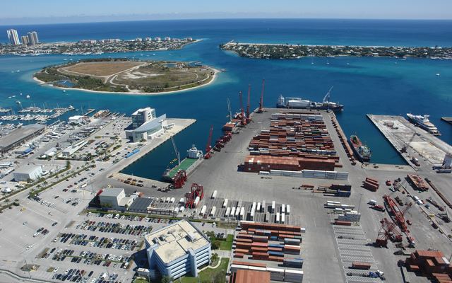 Aerial view east over the Port of Palm Beach in Riviera Beach, with stacked shipping containers and cranes in front, Peanut Island in the lagoon, and the Lake Worth Inlet between Singer Island and Palm Beach beyond