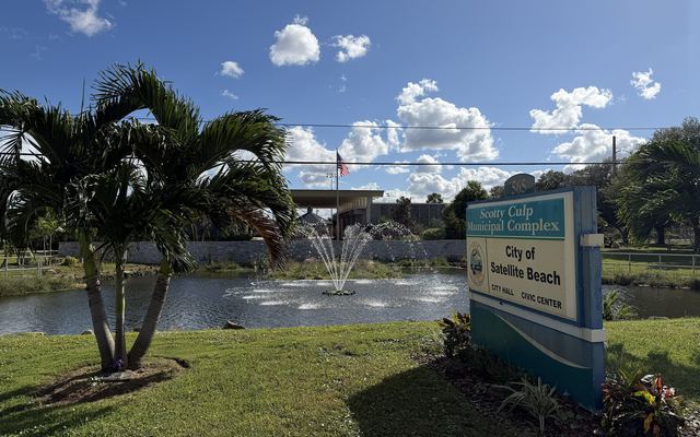 The Scotty Culp Municipal Complex sign for Satellite Beach City Hall and Civic Center beside a pond with a fountain, with palm trees, an American flag and the low city building under a blue sky
