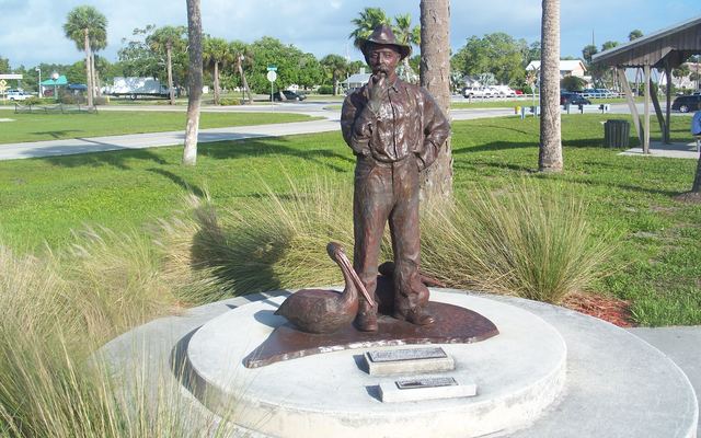 Bronze memorial statue of Paul Kroegel in a wide-brimmed hat, standing beside a bronze pelican on a round concrete base, set in grass and ornamental grasses in a Sebastian park with palm trees and a picnic pavilion