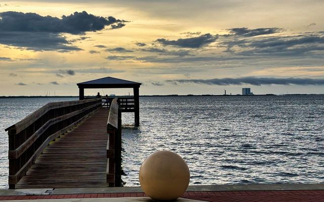A wooden fishing pier with a roofed shelter at its end reaching into the Indian River from the Space View Park waterfront in Titusville in the evening, with Kennedy Space Center's Vehicle Assembly Building small on the far shoreline