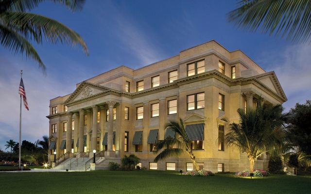 The restored 1916 Palm Beach County Courthouse in downtown West Palm Beach at dusk, its columned facade lit up behind a lawn, palm trees and an American flag